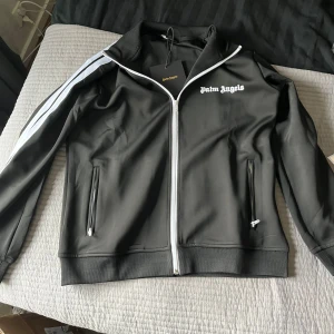 Svart track jacket från Palm Angels - Säljer en svart track jacket från Palm Angels med vit dragkedja och vita ränder längs ärmarna. Jackan har ståkrage, två fickor med dragkedja och logga broderad på bröstet. Perfekt för en sportig och trendig look.