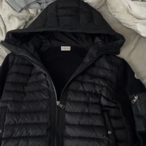 Moncler cardigan - Säljer en svart Moncler cardigan med huva och dragkedja. Jackan har quiltad framsida och stickade ärmar, samt Moncler-logga på ena ärmen. Perfekt för kyliga dagar🔥🔥🥰