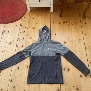 Grå vindjacka från Under Armour - Säljer en grå vindjacka från Under Armour med huva och dragkedja. Jackan har en stilren design med två nyanser av grått och det ikoniska logotyptrycket på bröstet. Perfekt för blåsiga dagar.