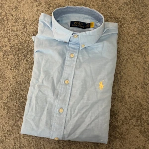 Ljusblå skjorta från Polo Ralph Lauren - Säljer en stilren ljusblå skjorta från Polo Ralph Lauren. Skjortan har långa ärmar och knappar framtill. Den är dekorerad med det klassiska broderade logotypen i gult på bröstet. Perfekt för en elegant look.