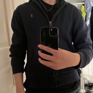 Mörkblå ziphoodie från Polo Ralph Lauren - Säljer en mörkblå hoodie från Polo Ralph Lauren med dragkedja och klassisk röd broderad logga på bröstet. Jackan har huva och fickor framtill, perfekt för en avslappnad stil. Väldigt skön inuti också med mjukt material.                                             NYPRIS: 1100kr.    STORLEK: S/M.               