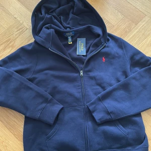 Mörkblå hoodie med dragkedja från Polo Ralph Lauren - Säljer en helt oanvänd mörkblå hoodie från Polo Ralph Lauren med dragkedja och huva. Jag köpte den men märkte att den var lite för stor för mig. Tröjan har en röd broderad logga på bröstet och två fickor framtill. Perfekt för en avslappnad stil. STORLEK: S/M.      NYPRIS: 1100kr