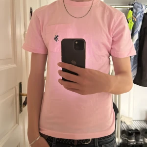 Ljusrosa t-shirt med bröstficka från U.S. Polo Assn. - Säljer en ljusrosa t-shirt från U.S. Polo Assn. med klassisk passform. T-shirten har en bröstficka med broderad logga i mörkblått. Perfekt basic-plagg med rund hals och korta ärmar.