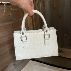 Vit handväska i krokopräglat mönster - Säljer en fin liten handväska, det ska finnas axelband till hittade inte som när jag skulle fota… det får plats ungefär en plånbok och en mobil beroende på hur stor mobil man har…