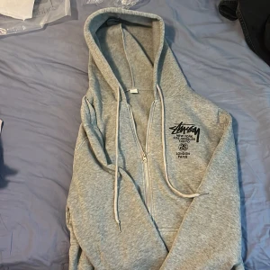 Grå zip-hoodie från Stüssy - Bara serösa köpare!!!  Säljer en grå hoodie med dragkedja från Stüssy. Tröjan har huva med dragsko och tryck både fram och bak med olika städer. Perfekt för en avslappnad streetwear-look.