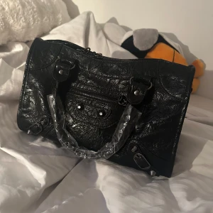 Svart handväska  - Säljer en svart handväska i skinn med snygga metalldetaljer och dragkedja upptill. Väskan har ett klassiskt format med korta handtag och en liten ficka framtill. Perfekt för dig som vill ha en stilren accessoar till din outfit. Den är helt ny aldrig använd då jag har en exakt likadan fast mini och köpt från en marknad i Spanien.❤️❤️