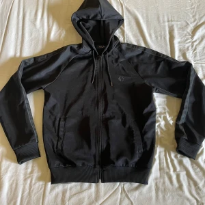 Svart hoodie från Fred Perry - Säljer en svart hoodiejacka från Fred Perry med dragkedja och fickor framtill. Använd ksk 3 gånger.