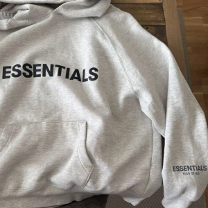 Essentials Fear of God - Säljer en ljusgrå hoodie från Essentials Fear of God. Tröjan har stor svart logga på bröstet och mindre logga på ärmen. Klassisk känguruficka framtill och huva. Perfekt för en avslappnad och trendig stil.