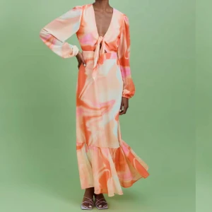 Färgglad långklänning med knyt och volang - Säljer en långklänning i mjuka orange, rosa och beige toner med abstrakt mönster. Klänningen har v-ringning, knyt framtill, långa puffärmar och volang nertill. Perfekt för sommarens tillfällen! Helt ny 
