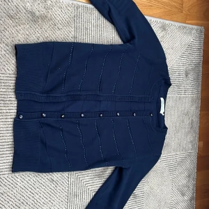 Mörkblå kofta med knappar - Snygg mörkblå kofta med knappar framtill och diskreta ränder. Perfekt för en stilren look. Koftan har långa ärmar och ribbade kanter för extra komfort. ❗️några diamanterna har ramlat av, men den märks inte så mycket❗️