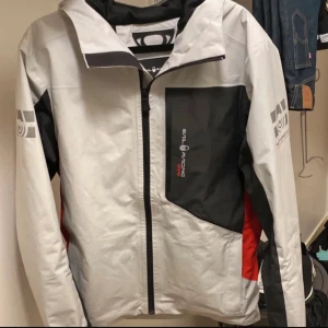Grå och svart vindjacka från Sail Racing - Säljer en grå vindjacka från Sail Racing med svarta och röda detaljer. Jackan har dragkedja framtill, hög krage och logga på bröstet samt ärmarna. Perfekt för blåsiga dagar och snygg sportig look.