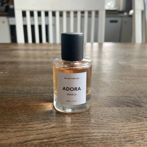 ADORA ANGELIA Eau de Parfum - En stilren parfymflaska med svart lock och transparent glas som visar en ljus bärnstensfärgad vätska. Märkt med ADORA ANGELIA, 50 ml Eau de Parfum. Flaskan har en minimalistisk design och passar perfekt på parfymhyllan. Denna lukt är inspirerad av Angela Share.