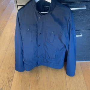 Mörkblå jacka från J.Lindeberg - Säljer en stilren mörkblå jacka från J.Lindeberg med hög krage och flera fickor framtill. Jackan har knappar och dragkedja samt lång ärm. Perfekt för dig som gillar klassisk och funktionell design.