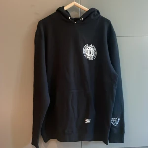 Svart hoodie från Diamond Supply Co. - Säljer en svart hoodie från Diamond Supply Co. med Grizzly Griptape-logga på bröstet och ryggen. Tröjan har en stor ficka framtill och en snygg diamantdetalj på ärmen. Perfekt för en avslappnad stil.