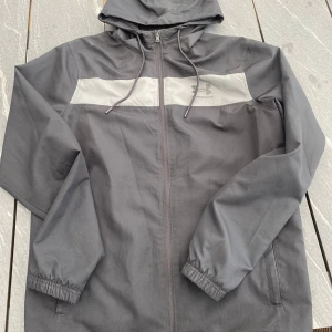 Grå vindjacka från Under Armour - Säljer en grå vindjacka från Under Armour med huva och dragkedja. Jackan har en ljusgrå panel med logga över bröstet, elastiska muddar vid ärmslut och meshfoder på insidan. Perfekt för utomhusaktiviteter.