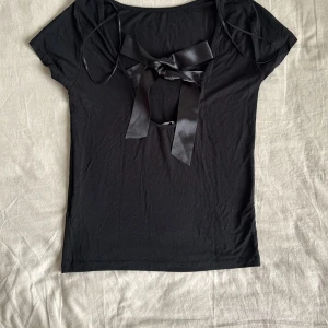 Svart topp med rosett - Snygg svart topp med korta ärmar och en stor satinrosett framtill. Toppen har en djupare urringning och är tillverkad i ett mjukt material som sitter skönt på kroppen. Perfekt för att lyfta en enkel outfit.