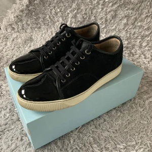 Lanvin - Säljer ett par svarta sneakers från Lanvin med snygga lackade detaljer på tån och häl. Skorna har snörning, vit sula och är tillverkade i en mix av mocka och skinn. Perfekta för dig som vill ha en stilren och lyxig look. 10/10 skick. Passar strl 42-44