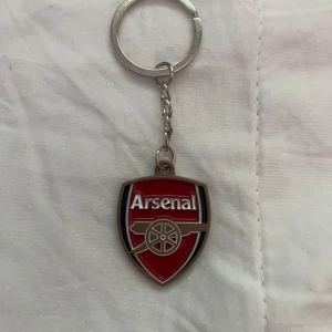 Arsenal nyckelring - Säljer en snygg nyckelring med Arsenal-loggan i rött, vitt och blått. Perfekt för dig som är ett fan av fotbollslaget Arsenal och vill visa ditt stöd på ett stilrent sätt.