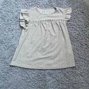 Beige blus från SHEIN med volangärmar - Söt vit blus med broderade detaljer och volangärmar. Blusen har rund halsringning och ett diskret hålmönster över hela tyget. Perfekt för en fräsch och somrig look. Pris kan diskuteras!