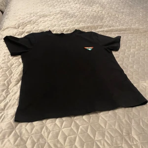 Svart t-shirt från Prada - Säljer en svart t-shirt från Prada med klassisk passform och korta ärmar. På bröstet finns Pradas ikoniska trekantslogga i rött, grönt och vitt. Perfekt för dig som gillar stilrena designer och exklusiva märken.