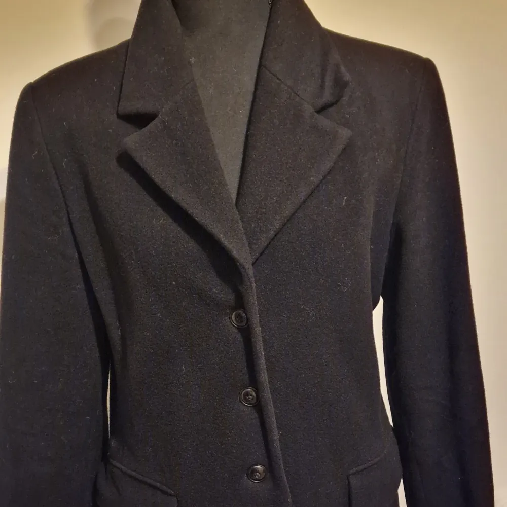 Stilren svart kappa i ull med klassisk krage och tre knappar framtill. Kappan har långa ärmar och två fickor med lock. Perfekt för kyligare dagar och ger en tidlös look.. Takit.