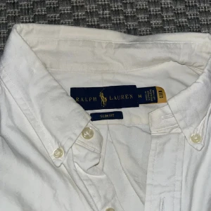 Vit skjorta från Ralph Lauren - Säljer en klassisk vit skjorta från Ralph Lauren i slim fit. Skjortan har långa ärmar och knappar framtill samt en broderad logotyp på bröstet. Perfekt för en stilren look.
