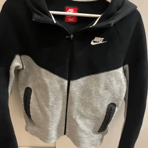 Nike tech fleece  - Säljer en snygg svart och grå hoodie från Nike med dragkedja och huva. Hoodien har en klassisk design med Nike-logga på bröstet och praktiska fickor framtill. Perfekt för en sportig look! Nypris: 1100kr. Enda slitaget är några få trådar på framsidan.