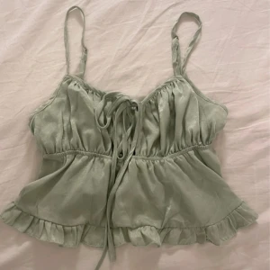 Grön satinlinne med volangdetaljer - Säljer ett sött grönt satinlinne med smala axelband och volangdetaljer i nederkant. Toppen har en knytning framtill som ger en fin passform. Perfekt för varma dagar! Aldrig använd!!