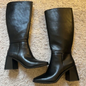 Svarta höga stövlar med klack - Säljer ett par svarta höga stövlar med bred klack och rund tå. Skorna är i skinnimitation och har en stilren design som passar till många outfits. Perfekta för dig som vill ha ett par klassiska boots med lite höjd.