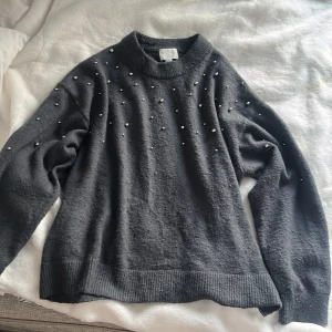 Mörkgrå stickad tröja med pärlor från H&M - Säljer en mörkgrå stickad tröja från H&M med dekorativa pärlor på axlar och överdel. Tröjan har lång ärm, rund halsringning och breda ribbade muddar vid ärmslut och nederkant. Perfekt för dig med Stockholmstil. 