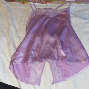 Lila siden nattlinne med spetsdetaljer - Elegant lila nattlinne i siden med vackra spetsdetaljer. Axelbandslös design med en liten rosett framtill. Matchande shorts med elastisk midja och spetskant. Perfekt för en lyxig natt. sparsamt använd..