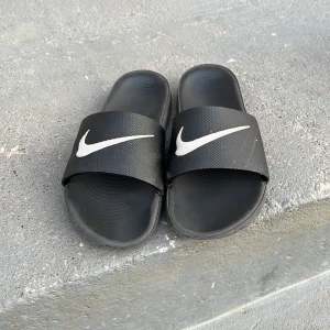 Svarta badtofflor från Nike - Säljer ett par svarta tofflor från Nike med den klassiska loggan i vitt på ovansidan. Perfekta för stranden eller poolen. De har en bekväm passform.