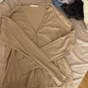 Beige kofta från Wera Stockholm - Säljer en stilren beige kofta från Wera Stockholm. Koftan har en v-ringad design med knappar framtill och långa ärmar. Perfekt för lager-på-lager under kyligare dagar.