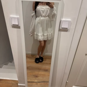 Vit offshoulder klänning med volang - Charmig vit offshoulder klänning med prickigt mönster och volangdetaljer. Klänningen har en knytning i midjan och långa ärmar. Perfekt till studenten!
