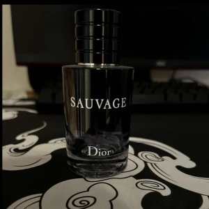 Sauvage parfym från Dior - Elegant parfymflaska från Dior med namnet Sauvage. Flaskan är mörk och stilren med en svart kork och vit text. Perfekt för den som söker en sofistikerad doft.