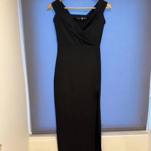 Svart långklänning från Boohoo - Elegant svart långklänning från Boohoo med omlott-design och ärmlös stil samt en slits framtill. Perfekt för en stilren look.