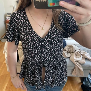 Svart blommig blus från Monki - Säljer en söt svart blus från Monki med rosa och vita blommor. Blusen har en v-ringning och knappar framtill samt volangdetaljer vid ärmarna. Perfekt för en somrig look! 🌸