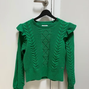 Fin grön stickad tröja från Holly & Whyte, Lindex. Strl XS - Snygg grön stickad tröja från Lindex med flätmönster och volangdetaljer vid axlarna. Perfekt för att hålla sig varm och stilren under kyligare dagar. Tröjan har långa ärmar och en rund halsringning.