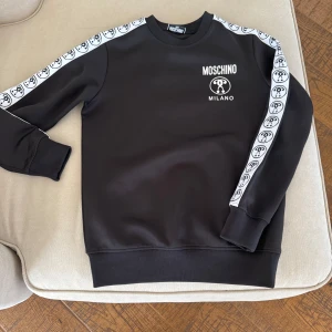 Svart sweatshirt från Moschino - Snygg svart sweatshirt från Moschino med vita detaljer. Tröjan har långa ärmar med ett mönster av logotyper längs ärmarna och Moschino Milano-tryck på bröstet. Perfekt för en stilren look. Jättefint skick. Använd två gånger. Pris går att diskutera. Storlek 164. 