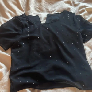Svart blus med bling bling från H&M - Säljer en elegant svart blus från H&M med små bling bling. Blusen har korta ärmar och en knappdetalj i nacken. Perfekt för en stilren look.