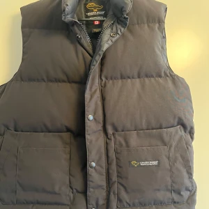 Svart dunväst från Canada Goose  - Svart dunväst från Canada Goose med hög krage. Västen har både dragkedja och tryckknappar. Använd få gånger 