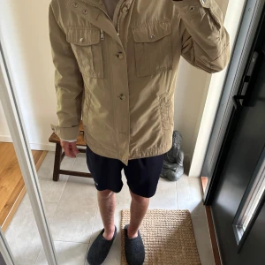 J Lindberg Fieldjacket (pris diskuteras)   - Snygg beige vindjacka med flera praktiska fickor framtill och både knappar och dragkedja. Jackan har en rak passform och lång ärm, perfekt för dig som gillar en clean och stilren look. Passar till många olika outfits och är lätt att matcha.