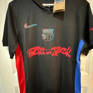 FC Barcelona x Nike Cactus Jack tröja - Snygg svart FC Barcelona fotbollströja från Nike med Cactus Jack-grafik i rött framtill. Tröjan har klubbmärke på bröstet, röda och blå paneler på sidorna och korta ärmar. Materialet är lätt och andas, perfekt för träning eller match.