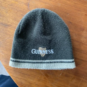 Guinness grön mössa med brodyr - Mörkgrön mössa från Guinness med broderad logga och harpa framtill. Två ljusare ränder nertill ger en snygg kontrast. Tillverkad i mjukt syntetmaterial - akryl - som håller dig varm. Perfekt accessoar för dig som gillar klassiska ölvarumärken.