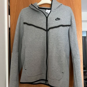 Nike tech fleece - Snygg grå Nike teech hoodie från Nike med svart logga på bröstet och hel dragkedja framtill. Tröjan har huva, svarta detaljer längs bröstet och en extra ficka med dragkedja på ena ärmen. 