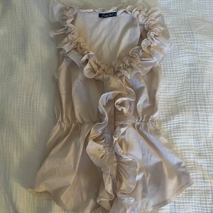 Beige ljusrosa volangblus  - Beige blus med volanger längs v-ringningen och framtill. Ärmlös modell med knappar framtill och markerad midja. 