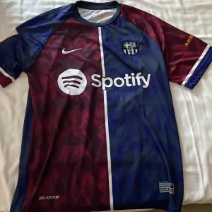 FC Barcelona Lamine Yamal Nike tröja - Snygg FC Barcelona matchtröja med Lamine Yamal 27 på ryggen. Tröjan är i Nike Dri-FIT material, har korta ärmar och är delad i vinröd och marinblå med vita detaljer. Sponsorer som Spotify och UNHCR tryckta på framsidan och baksidan. Perfekt för dig som älskar Barca!