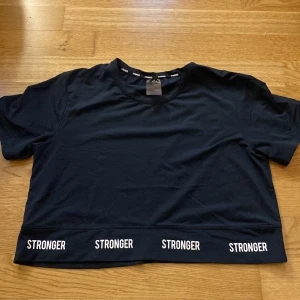 Svart croppad topp från Stronger - 💕Snygg svart croppad topp från Stronger med kort ärm och vit logga tryckt flera gånger längs nederkanten. 💕