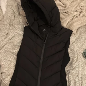Svart dunväst med huva från Zara - Snygg svart dunväst från Zara i storlek XS. Västen har en stor, vadderad huva och dragkedja framtill. Quiltad design med breda paneler och en clean look. Perfekt för lager-på-lager och streetstyle.