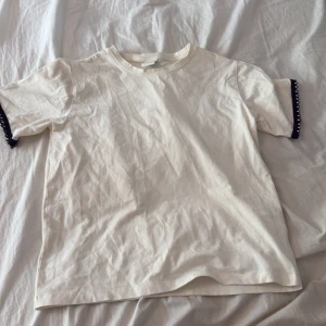 Vit t-shirt med pärldetaljer från Zara - Vit t-shirt från Zara i mjuk bomull med korta ärmar. Ärmsluten har svarta spetsdetaljer med små pärlor som ger en snygg kontrast. 